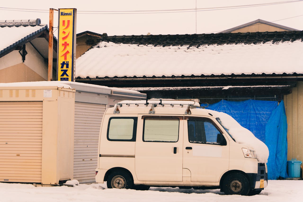 A small van covered in snow parked outside a traditional Japanese house during winter.
